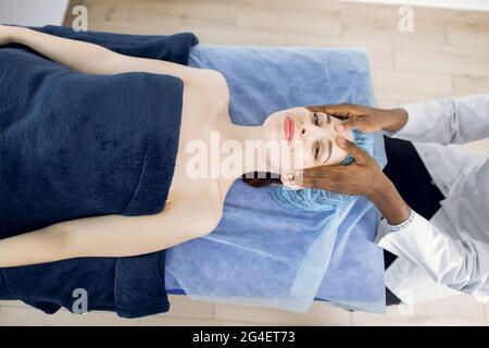 Top view of smiling pleasant relaxed young woman, enjoying facial massage in modern spa clinic. Hands of unrecognizable african male masseur massaging client's face and forehead. Stock Photo