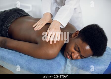 Close up of woman doctor physiotherapist manipulating shoulder blade on young African American man. Man laying on spa bed with doctor's hands massaging shoulder and back Stock Photo