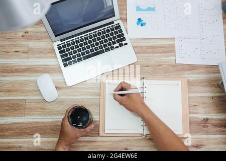 Entrepreneur drinking glass of fresh water and taking notes in planner after analzying sales reprot, view from the top Stock Photo