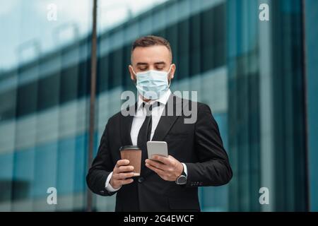 Male entrepreneur in suit wearing face mask and using smartphone, going to office and drinking morning coffee Stock Photo
