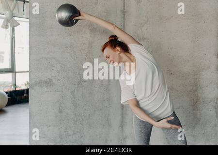 Athletic ginger haired young woman performing barre exercise with small fitball Stock Photo