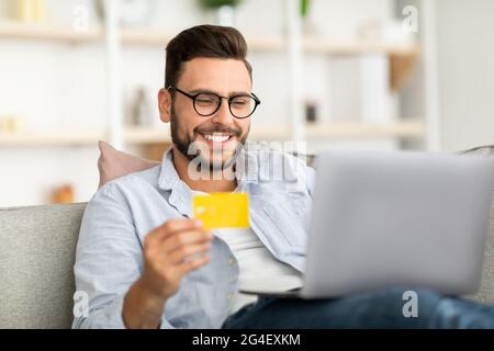 Easy payment and cashback concept. Smiling young man holding credit card and using laptop computer, sitting on couch Stock Photo