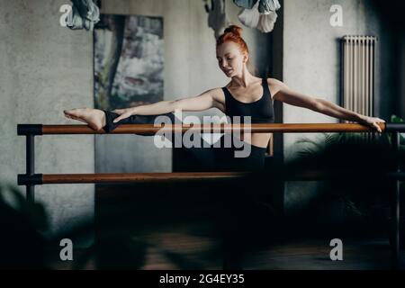 Young red haired woman in good body shape doing stretching exercises on ballet barre Stock Photo