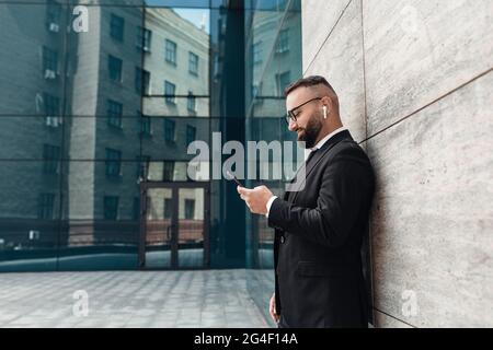 Entrepreneur listening to music in wireless earphones and using smartphone, standing outdoors, empty space, side view Stock Photo