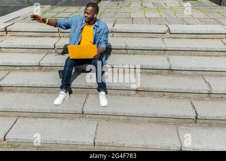 Joyful afro american freelancer businessman using yellow laptop outdoors,sitting on stairs in the park, talking online with colleagues or family Stock Photo
