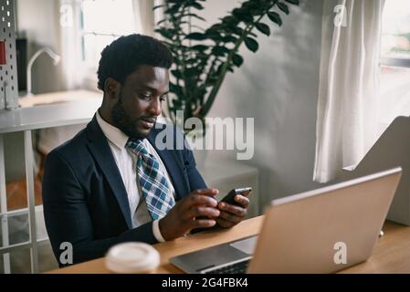 Black employee chatting on smartphone against laptop in office Stock Photo
