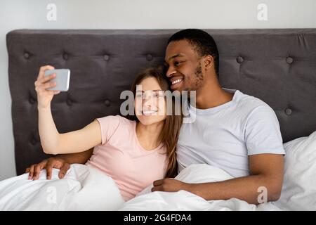 Millennial multiracial couple with smartphone taking selfie in bed before going to sleep, indoors Stock Photo