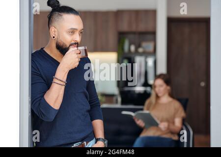 Portrait of pensive handsome man with his hair in bun standing at window and driking tea Stock Photo