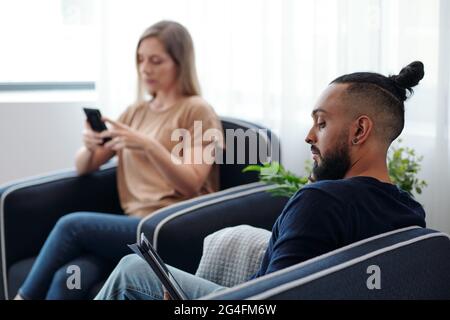 Concentrated bearded man reading e-book, his wife checking text messages on smartphone in background Stock Photo