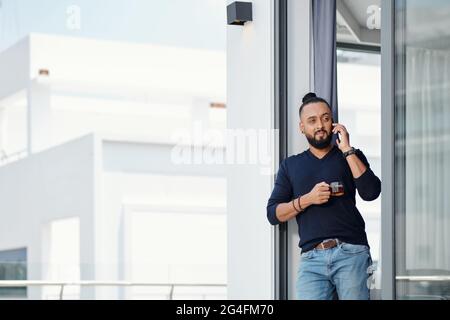 Pensive handsome bearded man drinking tea and talking on phone with friend or colleague Stock Photo