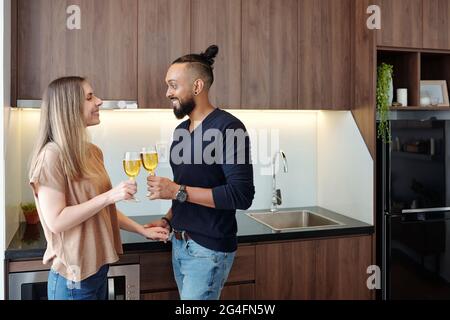 Smiling boyfriend and girlfriend with glasses of wine standing in kitchen and looking at each other, celebrating anniversary Stock Photo