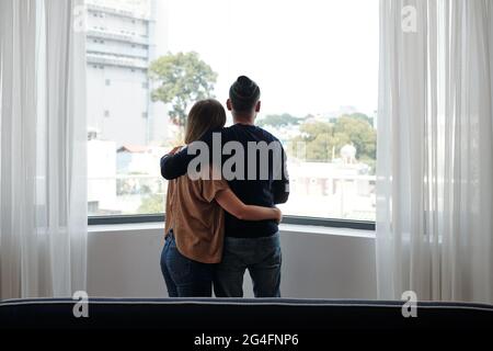 Hugging young couple standing at big window and looking outside, view from the back Stock Photo