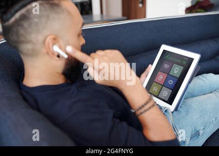 Man sitting on sofa and controlling temperature and light in apartment via application on tablet computer Stock Photo