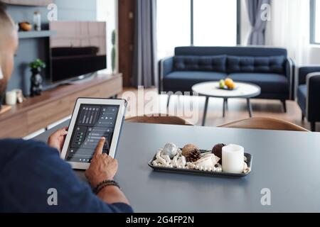 Man sitting at table and using smart home application on digital teblet when dimming lights in the room Stock Photo