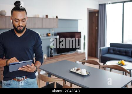 Man with wireless earbuds standing in his apartment and choosing music on tablet computer Stock Photo