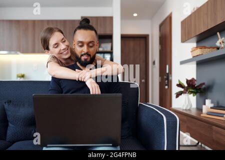 Happy young woman hugging her husband from behind when he is coding on laptop at home Stock Photo