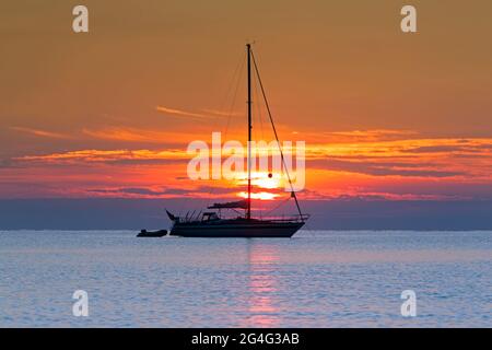 Sailing boat / sailboat / yacht with dinghy anchored with lowered sails, silhouetted against orange sky at sunset Stock Photo