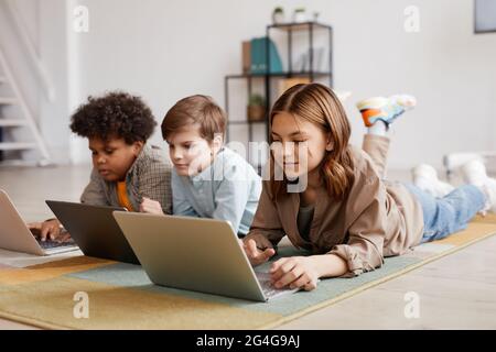 Full length view at group of three kids using computer devices while laying on floor, focus on smiling girl in foreground Stock Photo