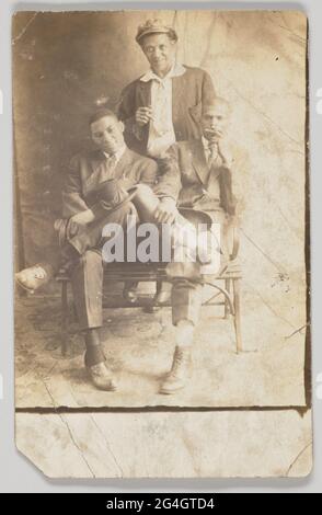 A photographic postcard of three men. The men appear to be posed on a backdrop in photography studio. Two men are seated on a bench and the third man stands behind them. The men are wearing suits and have hats. The seated men have their hats resting on their legs, while the standing man wears his hat. The standing man and the man seated on the right side of the image are holding cigarettes. All three men are facing the camera. The back of the photographic postcard is unused and has [POST CARD] printed in black at the top and blank spaces for [CORRESPONDENCE HERE] and [NAME AND ADDRESS HERE]. I Stock Photo
