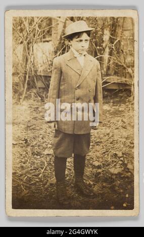 A photographic postcard of a young boy wearing a bowler hat, double-breasted jacket, breeches, and boots. He is standing outdoors on ground strewn with branches and fallen leaves. His body is turned slightly to the left, and he is looking away from the camera. The back of the photographic postcard is unused and has [POST CARD] printed in black at the top and blank spaces for [CORRESPONDENCE HERE] and [ADDRESS ONLY]. Stock Photo