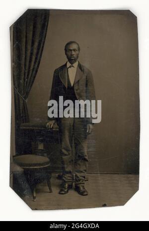 An unidentified African American man standing for his portrait in front of a drapery. He wears a jacket, waistcoat, and necktie, and a gold ring has been painted on to his right ring finger. Stock Photo