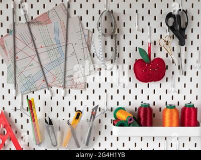 Pegboard with sewing supplies for tailor. White board for accessories storage. Sheets of clothing patterns, pincushion, threads, scissors, etc. Stock Photo