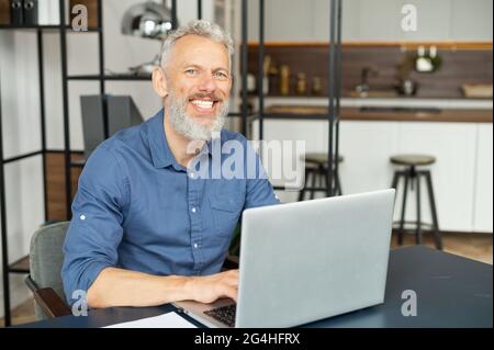 Portrait of mature grey-haired man using laptop in home office, senior male entrepreneur looks at the camera and smiling toothy, kitchen on the background. Elderly male freelancer Stock Photo