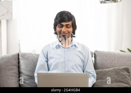 Cheerful optimistic young indian man wearing casual shirt using laptop, typing on keyboard, sending emails, chatting in social media, hindu freelancer guy working remotely sitting on the couch at home Stock Photo