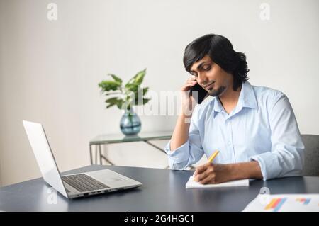 Intelligent indian male office employee talking on the smartphone and taking notes sitting at the desk in bright office space, using laptop computer, eastern man writes down during phone conversation Stock Photo