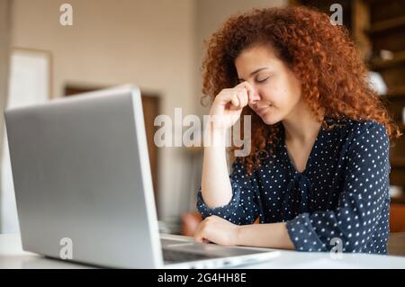 Upset and tired young curly red-haired woman sitting at the table at home using computer laptop with sad face expression, covering face with hands, feels depressed Stock Photo