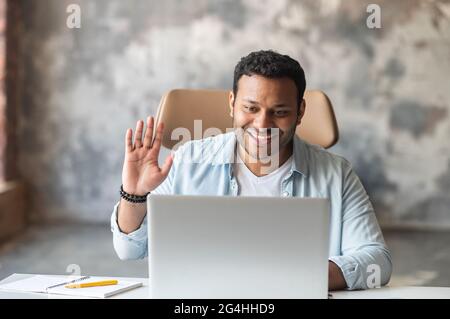 Morning meeting on the distance. Young indian male employee is using computer app on the laptop for video connection, guy taking a part in online webinar, waving into webcam and greeting participants Stock Photo