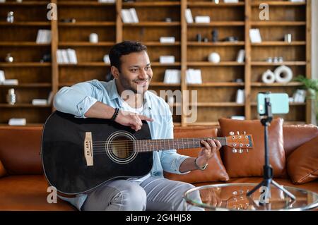 Handsome attractive indian guy making video blog about musical instruments, sitting on the couch, playing the guitar, and streaming tutorial online, using smartphone and tripod for recording Stock Photo