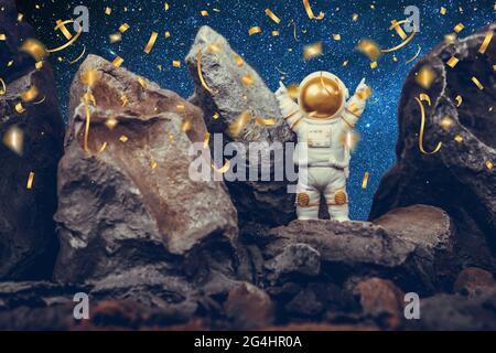 Astronaut standing on rocky mountain celebrating with raising arms while falling shiny golden confetti. Spaceman wearing white space suit and helmet. Stock Photo