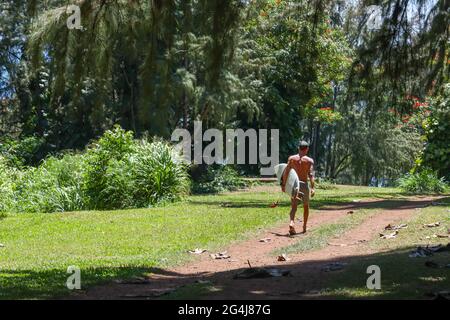 PRINCEVILLE, HAWAII, UNITED STATES - Jun 02, 2021: A male surfer carries his board down the walking trail at Kauai's Old Club Med Trail for quicker ac Stock Photo