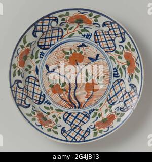 . Round plate of multi-colored padded faience, with a circle in the middle with multicolored flowers in it on a red shed behind. The edge has five vase-shaped compartments, each compartment is interspersed with a flower in the colors red, green and blue. Stock Photo