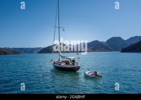 A sailboat underway with a man and woman on board motors out of Bahia Coyote in the Sea of Cortez with their dinghy under tow in calm blue water . Stock Photo