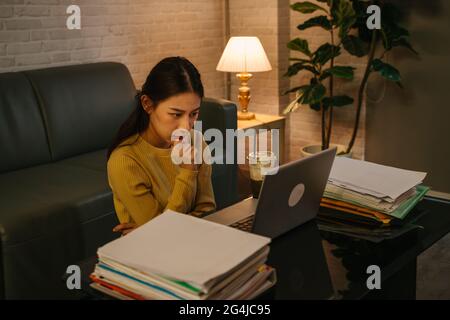 Tired young Asian female businesswoman thinking with hands on chin while staring at laptop screen with office working files on table during late night at home Stock Photo