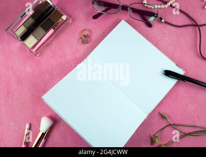 A large notebook in the center of the composition, women's cosmetics, makeup brushes lying on a pink background. Flat lay, top view. Stock Photo