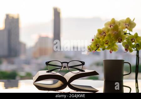 Spectacles put on open book with a cup of coffee, phalaenopsis orchid and city background. Stock Photo