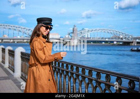 Happy young woman of Caucasian ethnicity in a light brown coat and black cap posing on the embankment against the backdrop of the bridge and the blue sky. Stock Photo