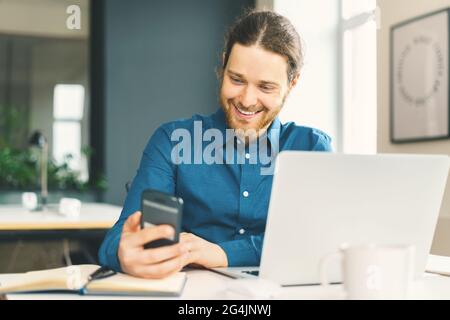 Smiling young man in casual wear looking at smartphone screen at workplace Stock Photo