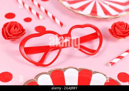 Red heart shaped glasses, surrounded by roses, confetti and party paper plates on pink background Stock Photo