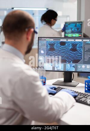 Man biologist checking DNA informations typing on pc in modern equipped laboratory. Group of biochemistry doctors examining virus evolution using high tech for researching treatment. Stock Photo