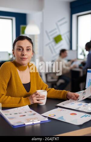 Businesswoman working with company charts looking at camera holding cup of coffee. Executive entrepreneur, manager leader sitting working on projects with diverse colleagues. Stock Photo