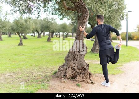 Young runner man with mask stretching in a park leaning against a tree Stock Photo