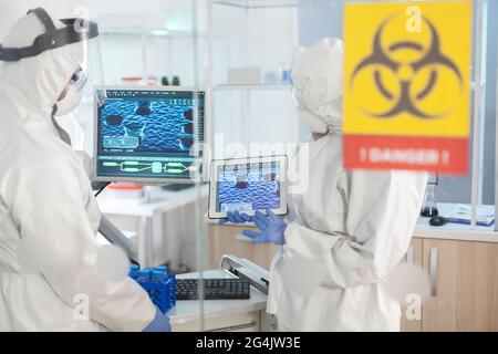 Doctors wearing ppe suit working in lab danger zone area using tablet pc. Group of doctors examining vaccine evolution using high tech for diagnosis against covid19. Stock Photo