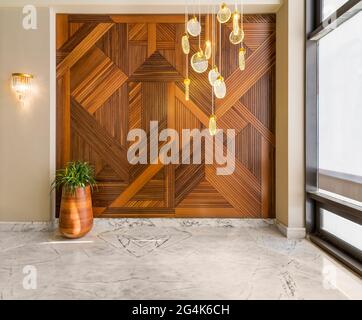 Hall with wood cladding wall decorated with geometrical patterns, tall rounded wooden planter with green bushes, contemporary glass tall chandelier, big window, and white marble floor in modern house Stock Photo