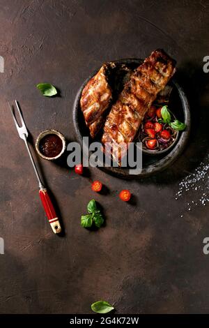 Grilled pork bbq ribs in ceramic plate served with meat fork, vegetable salad, cherry tomatoes, basil and barbeque sauce over dark brown texture backg Stock Photo