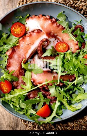 Seafood salad. Coocked tentacles of octopus on blue ceramic plate served with rocket leaf aragula and cherry tomato salad over brown wooden background Stock Photo