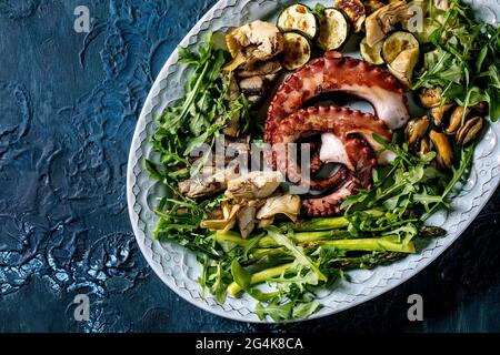 Seafood salad. Coocked grilled tentacles of octopus, sardines and mussels on blue ceramic plate with arugula salad, zucchini and asparagus over blue t Stock Photo